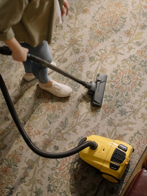 A person is vacuuming a decorative area rug with an ornate floral pattern featuring shades of beige, green, and muted orange in a well-lit indoor space. The individual is dressed in casual clothing, i